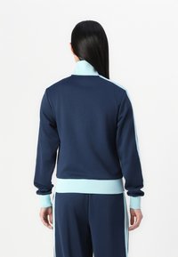 Mornarska trening jakna z svetlo modrimi detajli, rebrastimi manšetami in obodom, ki ima visok ovratnik in gladko teksturo. Model prikazan s hrbtne strani.