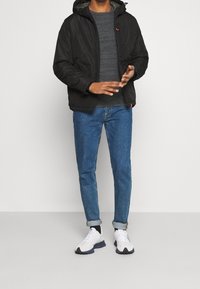 Giacca nera con cappuccio e chiusura a zip, maglione grigio a trama, jeans blu e sneakers bianche con dettagli grigi. Outfit casual con strati di texture.