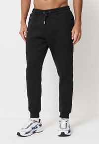 Pantalon de survêtement noir avec une taille à cordon, une coupe effilée et des poignets côtelés. Porté avec des baskets blanches et grises avec des accents bleus.