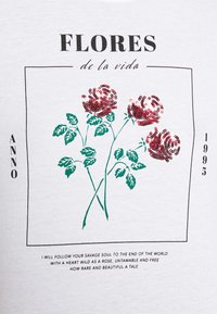 Hvid T-shirt med rød og grøn rose grafik med pailletter, sort tekst ovenfor og nedenfor, samt rektangulært ramme. Teksten inkluderer "Flores de la vida" og "Anno 1995."
