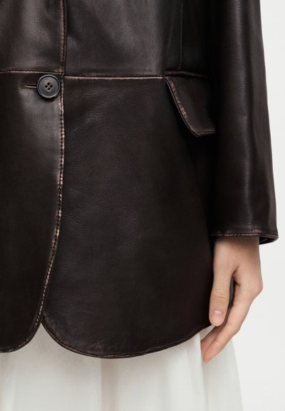 Καφέ σκούρο δερμάτινο μπουφάν με ένα μαύρο κουμπί, τσέπη με καπάκι, εμφανείς ραφές και ένα χέρι να ακουμπάει στο πλάι πάνω από λευκό ρούχο.