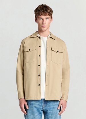 Beige button-up shirt with a collar, two chest pockets, and black buttons. Worn over a white crewneck shirt, paired with blue jeans.