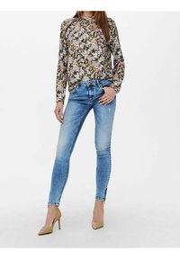 Bluse mit langen Ärmeln und floralen Mehrfarbmustern, kombiniert mit hellblauen Skinny-Jeans mit ausgewaschenen Stellen und einem Reißverschlussdetail an den Knöcheln.