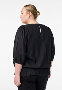 Melnā blūze ar pūkainām piedurknēm, struktūras audums, atslēgas formas izgriezums aizmugurē un savilktas maliņas. Apvienota ar melnu biksēm.