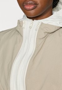 Ελαφρύ μπεζ μπουφάν με λευκή εσωτερική επένδυση και αντίθετο διπλό φερμουάρ. Διαθέτει ψηλό γιακά και υφή υλικού.