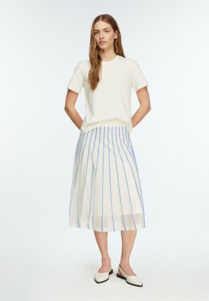 Kobieta stoi z rękami za plecami, ubrana w biały top z krótkim rękawem, białą spódnicę midi z niebieskimi pionowymi paskami oraz białe buty na niskim obcasie.