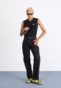 Black sleeveless cable-knit top with embroidered detail, high-waisted black jeans, and multicolored chunky sneakers with a green accent.