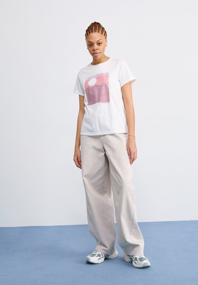 Weißes T-Shirt mit pinkem Grafikdruck, das Berge und eine Sonne zeigt. Kombiniert mit lockeren beigen Hosen und grau-weißen Sneakers. Heller Hintergrund.