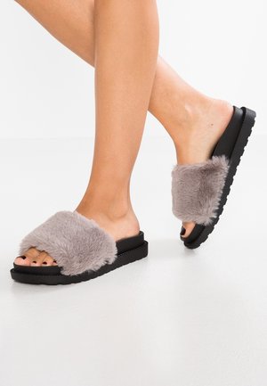 Osoba w czarnych klapkach na stopę z grubymi szarymi paskami z sztucznego futra, widoczne palce, na gładkiej, odbijającej białej podłodze.