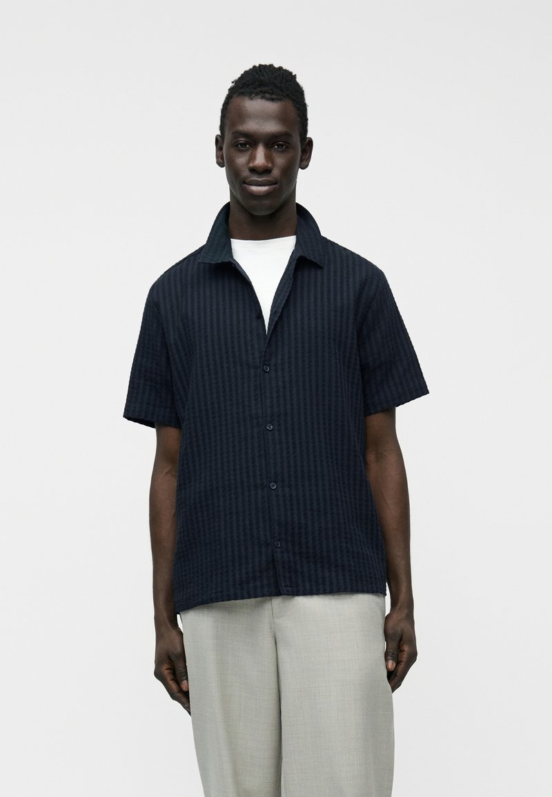 Homme portant une chemise à manches courtes bleu marine foncé boutonnée sur un t-shirt blanc, avec un pantalon gris clair, se tenant devant un fond clair uni.