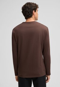 Langärmliges Hemd aus dunkelbrauner Baumwolle mit rundem Halsausschnitt und glatter Textur. Mit gesäumtem Saum und dezentem Branding in der Nähe des Saums.