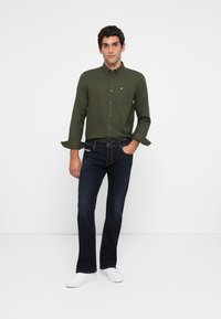 Hombre vestido con una camisa de botones verde oliva, jeans azul oscuro y zapatillas blancas, de pie contra un fondo blanco con una mano en el bolsillo.