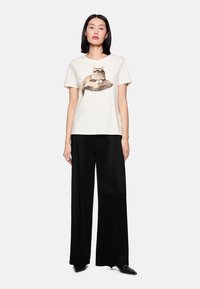 Baumwoll-T-Shirt in Creme mit einem Katzen-Motiv, kombiniert mit lockeren schwarzen Hosen. Das Design zeichnet sich durch eine entspannte Passform und kurze Ärmel aus.