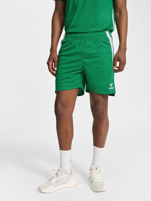 Mężczyzna w zielonych sportowych szortach z białymi bocznymi paskami, białych skarpetach i białych butach sportowych, stojący na jednolitym tle.