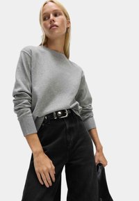 Jasnoszary sweter z okrągłym dekoltem i prążkowanymi mankietami, zestawiony z czarnymi spodniami o wysokim stanie i paskiem z srebrną klamrą.
