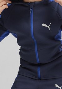 Giacca a zip navy con accenti blu, texture liscia, tasche con zip e logo Puma riflettente sul petto.