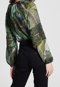 Nainen, jolla on ylleen vedetty läpinäkyvä vihreä paita, jossa on lehtikuviot ja levenevät hihat, yhdistettynä korkeavyötäröisiin mustiin farkkuihin, näkyy takaa.