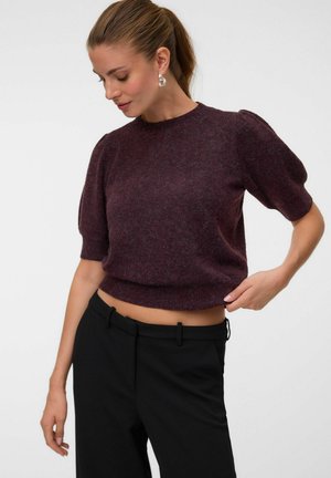Femme avec queue de cheval brune portant un pull violet à manches courtes et un pantalon noir regarde vers le bas, ajustant l'ourlet du pull de sa main droite.