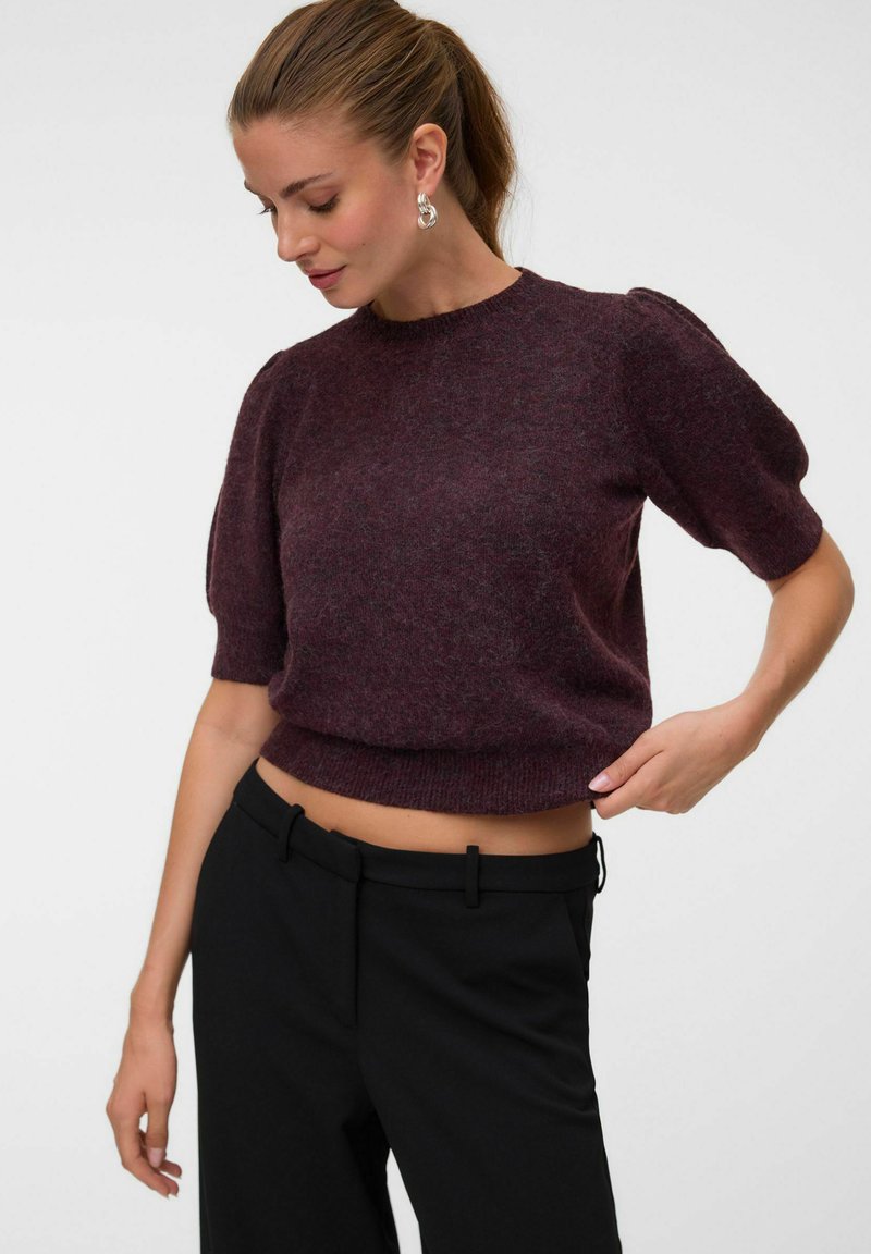 Femme avec queue de cheval brune portant un pull violet à manches courtes et un pantalon noir regarde vers le bas, ajustant l'ourlet du pull de sa main droite.
