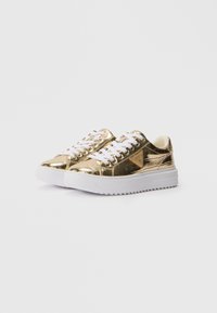 Adidași cu vârf jos, cu finisaj metalic auriu, șireturi albe, talpă groasă albă cu striații și logo triunghiular GUESS pe lateral.