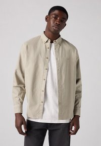 Lichtbeige blouse met lange mouwen en knoopsluiting, voorzien van een spitse kraag, met beige knopen en een textuurstof, gelaagd over een witte T-shirt.