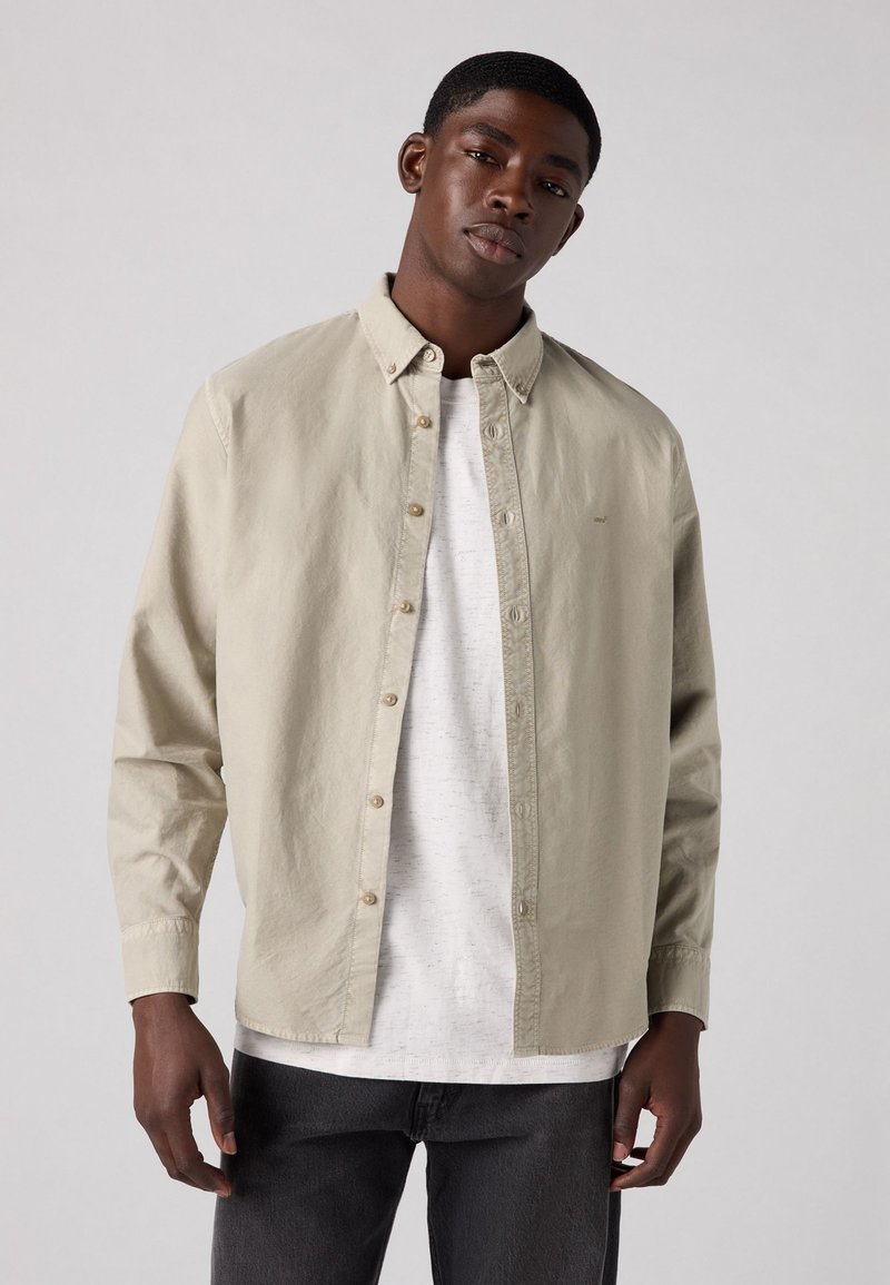 Lichtbeige blouse met lange mouwen en knoopsluiting, voorzien van een spitse kraag, met beige knopen en een textuurstof, gelaagd over een witte T-shirt.