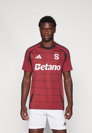 Weinrotes Fußballtrikot mit schwarzen Akzenten und horizontalen Streifen. Verfügt über das "S"-Logo und das Branding "Betano". Hergestellt aus atmungsaktivem Material.