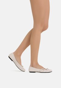 Scarpe piatte rosa chiaro con punta tonda, dotate di un dettaglio a fiocco annodato e di un tacco piccolo e solido nero. Texture liscia e design minimale.