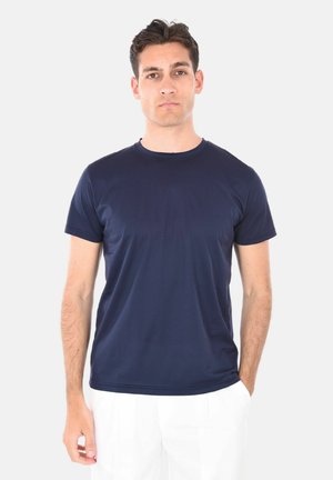 Giovane uomo con capelli corti e scuri indossa una maglietta a maniche corte blu navy e pantaloni bianchi, in piedi contro uno sfondo chiaro e uniforme.