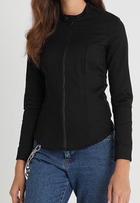 Svart figurnära jacka med hög krage och framdragkedja. Tillverkad av ett slätt material, med långa ärmar och rundad nederkant.