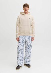 Hoodie beige avec poche avant et petit logo ; associé à un pantalon cargo blanc avec un motif marbré bleu ; chaussures marron avec semelles claires.