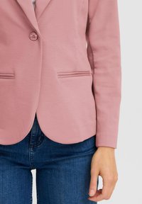 Blush roze blazer met een op maat gemaakt ontwerp, enkele knoopsluiting en twee voorzakken, gecombineerd met donkerblauwe jeans. Glad textuur.
