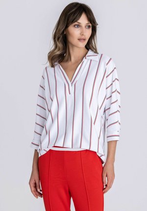 Femme portant un chemisier blanc à fines rayures verticales noires et rouges et un pantalon rouge taille haute, debout devant un fond clair uni.