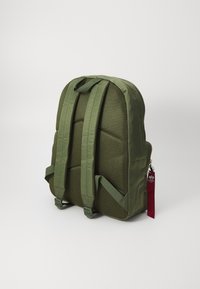Mochila verde oliva con respaldo de malla acolchada, correas ajustables, base plana, cierre de cremallera y una etiqueta de tela roja con detalles del logo.