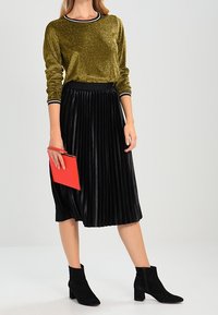 Frau trägt ein goldenes Langarmoberteil, einen schwarzen plissierten Midi-Rock, schwarze Stiefeletten und hält eine rote Clutch.