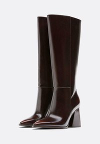 Ciemnobrązowe kozaki do kolan wykonane z błyszczącej skóry. Posiadają spiczasty nosek i masywny kwadratowy obcas z minimalnymi detalami szycia.