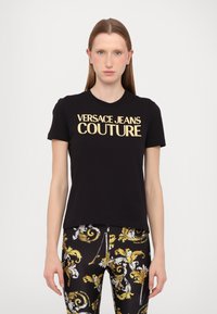 Juodas medvilnės marškinėliai trumpomis rankovėmis su ryškiu auksiniu "Versace Jeans Couture" logotipu krūtinėje.