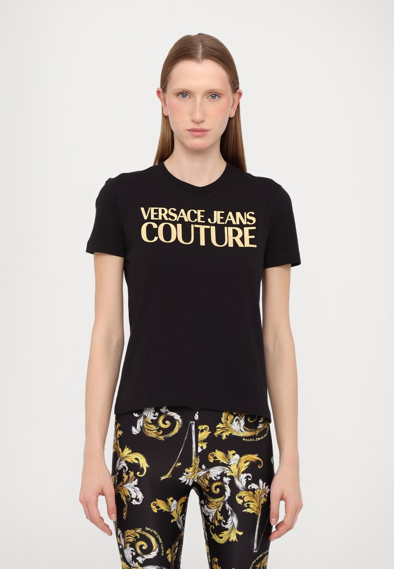Juodas medvilnės marškinėliai trumpomis rankovėmis su ryškiu auksiniu "Versace Jeans Couture" logotipu krūtinėje.
