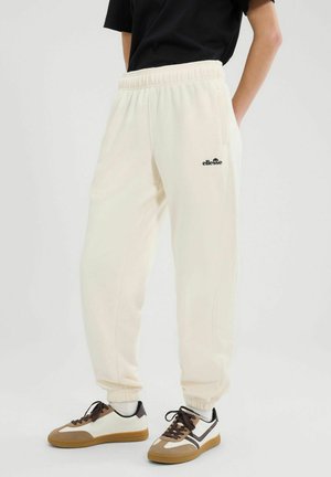 Persona che indossa pantaloni jogger color avorio con polsini elastici e logo nero, abbinati a sneakers stringate bianche, marroni e nere.