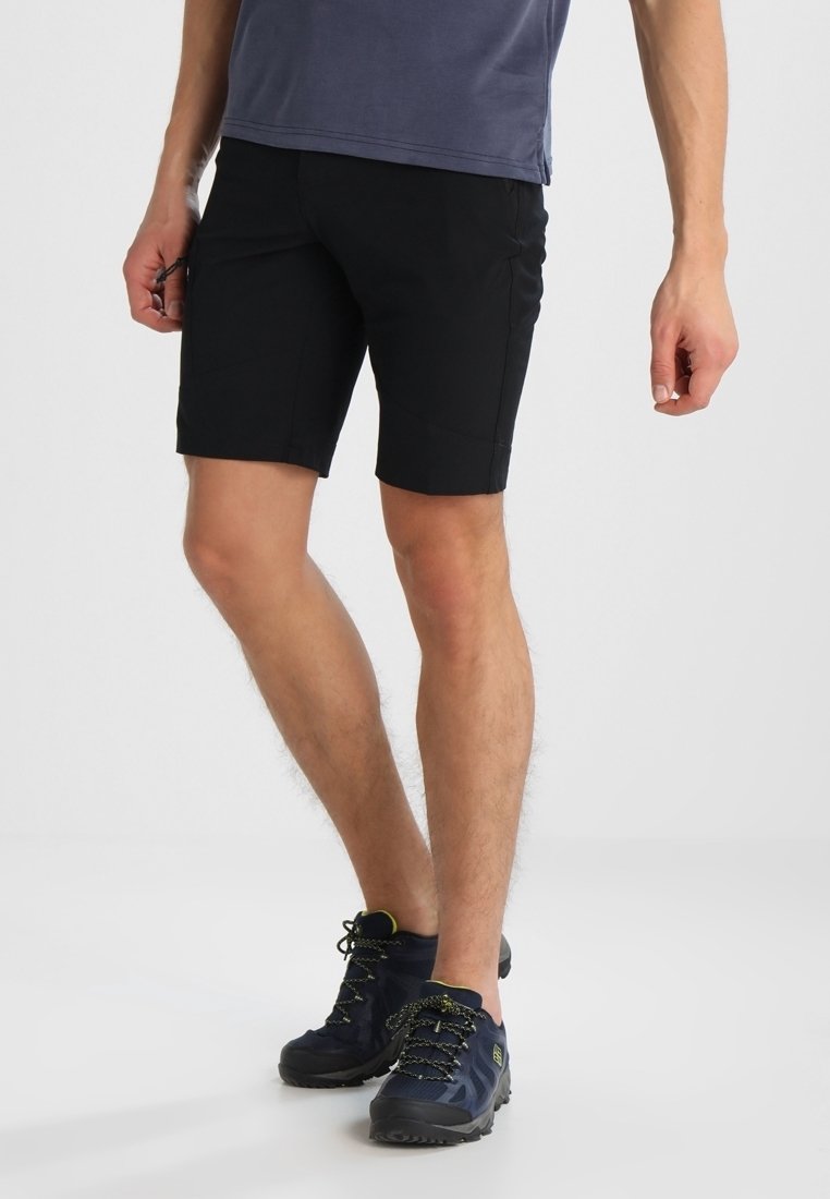 Homme portant un short noir, une chemise bleu marine et des chaussures de randonnée noires, debout les jambes croisées sur fond blanc.