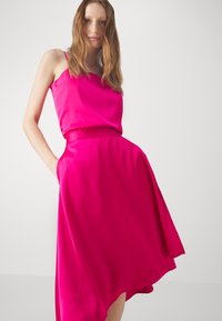Satin rosa kjole med ermeløs topp og asymmetrisk kant. Har en bred liv, samt sidelommer, som gir en flytende silhuett.