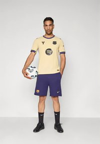 Beżowa koszulka piłkarska z czarnymi akcentami i logo, zestawiona z granatowymi spodenkami i czarnymi skarpetami. Model trzyma piłkę nożną, stojąc na neutralnym tle.