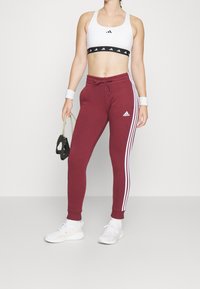 Bílá sportovní podprsenka s černými akcenty a logem, vínové tepláky s bílými pruhy po stranách a bílé tenisky, držící černý předmět.