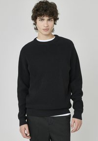 Un pull en maille côtelée noire avec un col rond et des manches longues, superposé sur une chemise blanche. Design simple et décontracté.