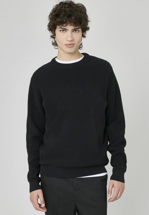 Un pull en maille côtelée noire avec un col rond et des manches longues, superposé sur une chemise blanche. Design simple et décontracté.