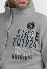 Graue Zip-Jacke mit hohem Kragen, versehen mit gesticktem Text und einem runden Abzeichen. Text in Marineblau, Weiß und orangefarbigen Akzenten. Weiche Textur.