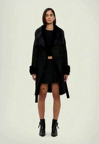 Schwarzer Mantel mit Kunstpelzkragen, langen Ärmeln und strukturierter Oberfläche. Kombiniert mit einem schwarzen Rock und Stiefeletten. Minimalistisches Design.