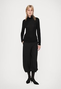 Crna turtleneck pulover, prilagođenog dizajna, uparen s labavim crnim skraćenim hlačama. Model nosi prozirne crne tajice i cipele sa potpeticom.
