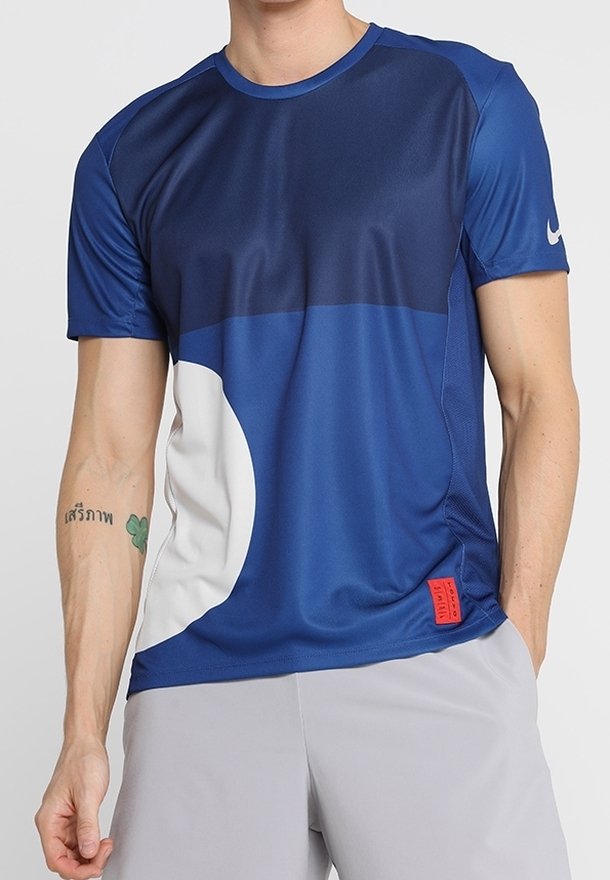 Homme portant un maillot de sport à manches courtes bleu et bleu marine avec des accents blancs et rouges et un short gris clair, debout avec une main dans la poche.