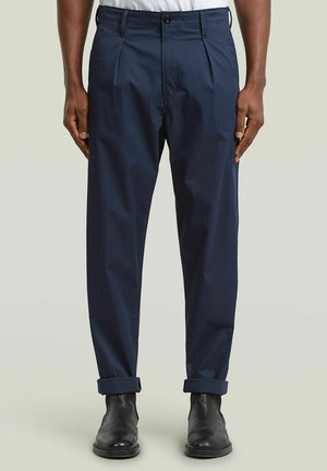 Homme portant un pantalon plissé bleu marine avec des revers roulés et des bottes en cuir noires, debout devant un fond clair uni.